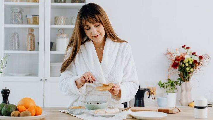 A woman in a bathrobe preparing breakfast in a home kitchen.